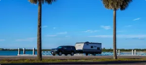 An SUV pulls a Coleman trailer along a road with a coastal waterway in the background.
